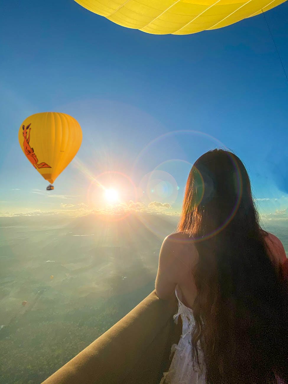 A person with long hair looking at a yellow hot air balloon and a sunrise while standing in a balcony.