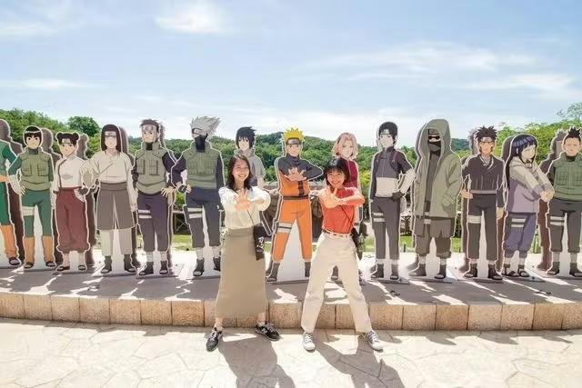 Two individuals posing in front of life-size cutouts of anime characters outdoors on a sunny day.