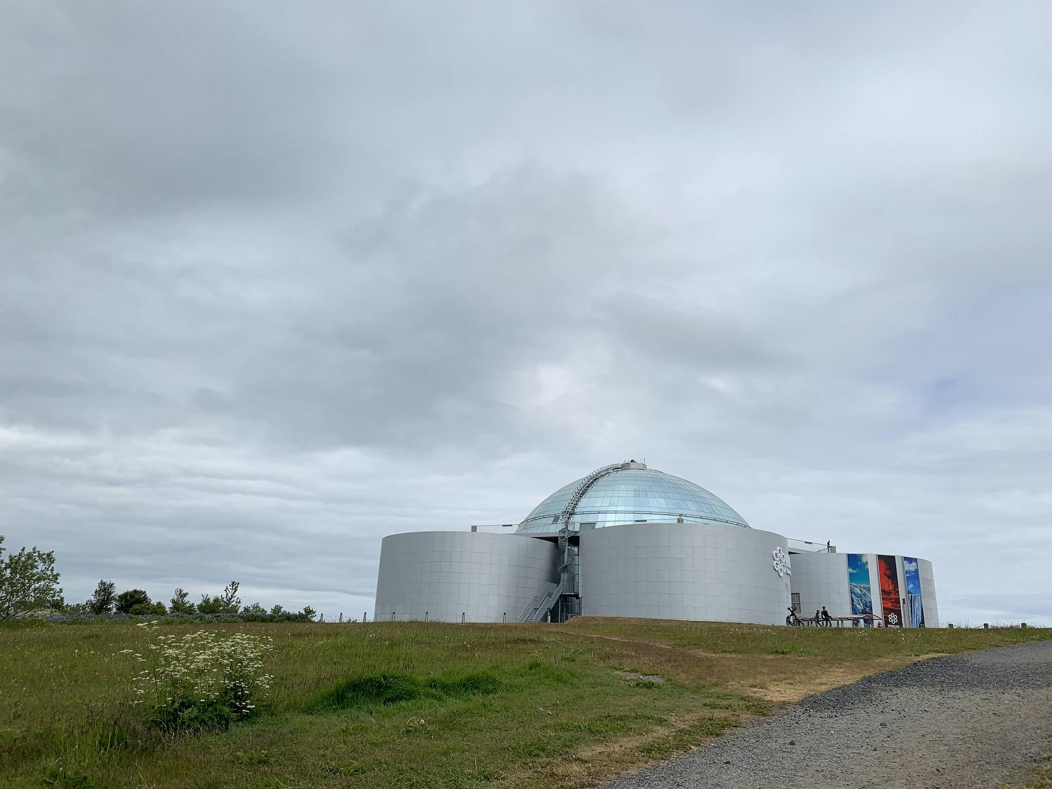 Un bâtiment cylindrique moderne avec un dôme en verre se trouve sur une colline herbeuse sous un ciel nuageux, avec des affiches sur le côté et des arbres à proximité.