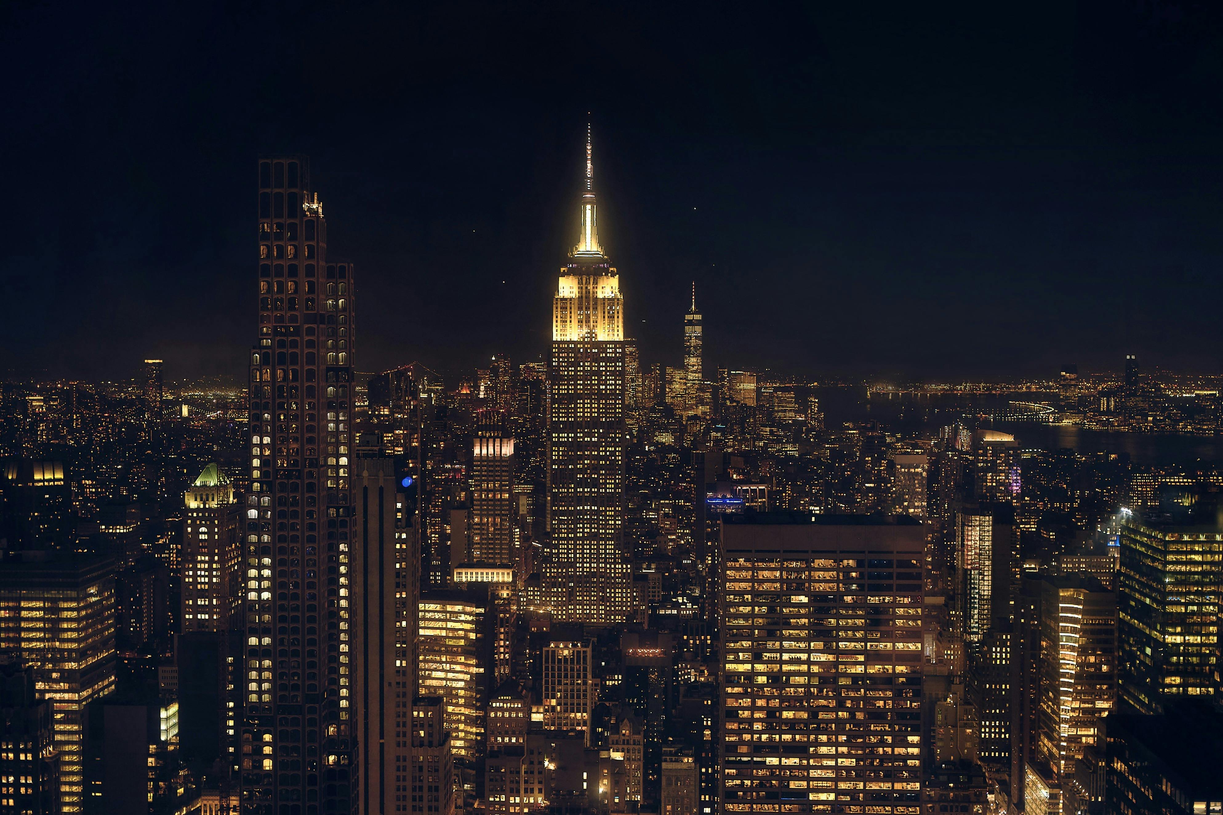 A nighttime cityscape featuring the illuminated Empire State Building surrounded by other lit-up skyscrapers.