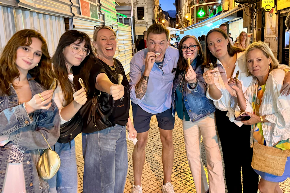 A group of six people smiling and holding small food items, standing on a cobbled street with buildings in the background at dusk.