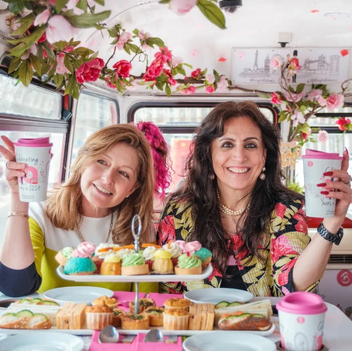 Two women smiling, holding cups, sitting in a decorated van with flowers and pastries on a table.