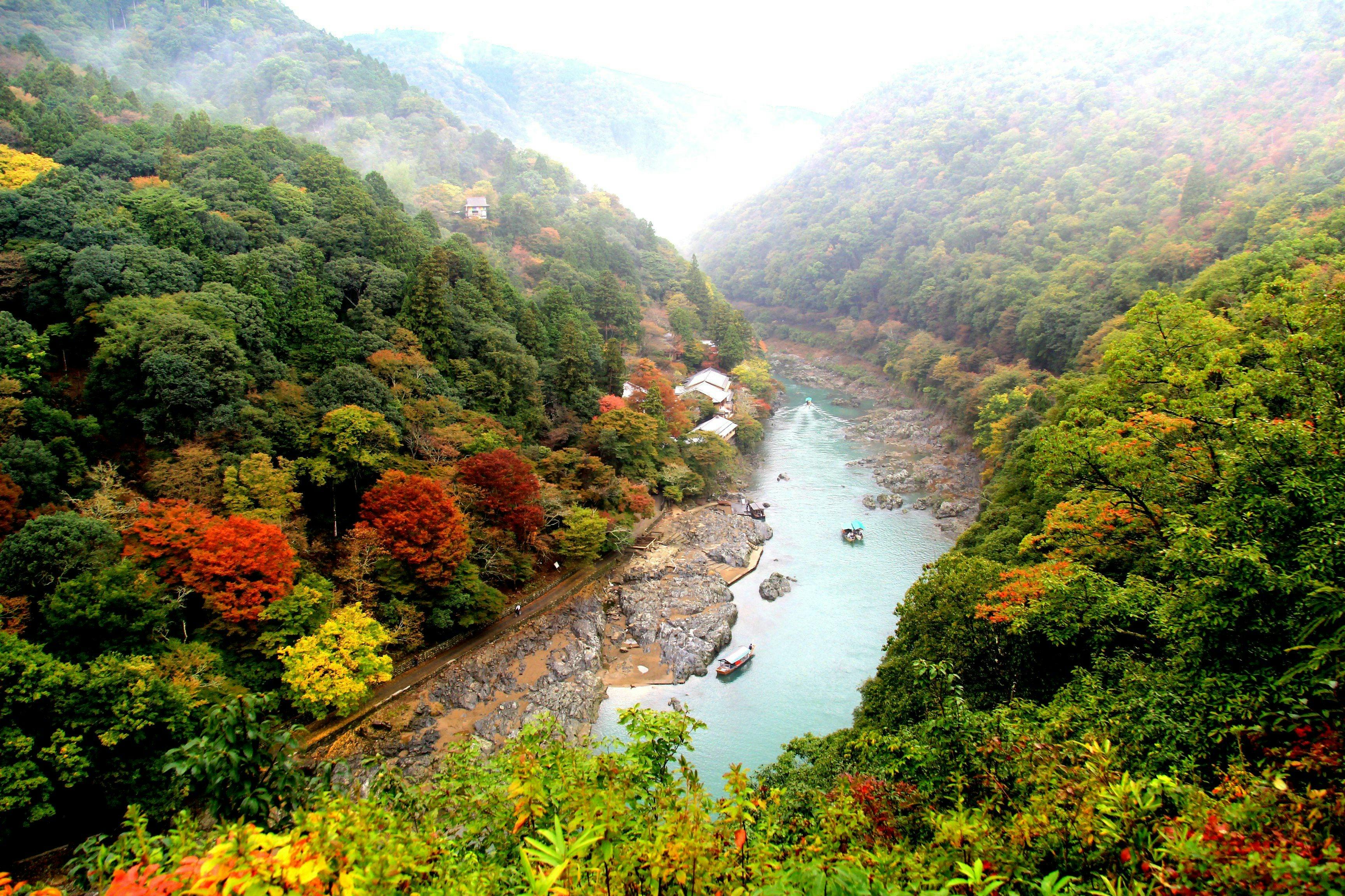 A river winding through a lush valley with dense, colorful forested hills and small buildings along the riverbank.