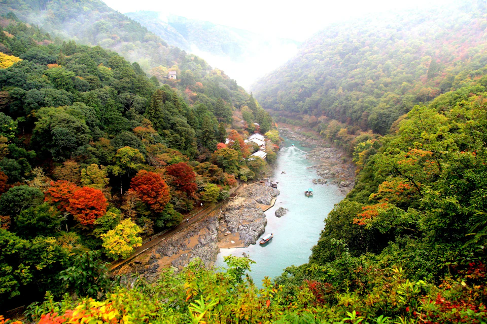 A river winding through a lush valley with dense, colorful forested hills and small buildings along the riverbank.