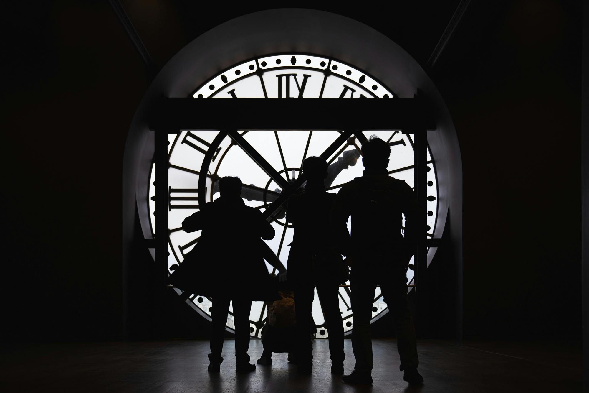 Three silhouetted figures stand in front of a large backlit clock face with Roman numerals.