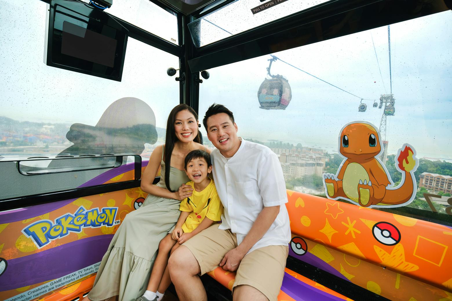 A family of three smiling inside a themed cable car cabin with cartoon decorations. A cable car is visible in the background.