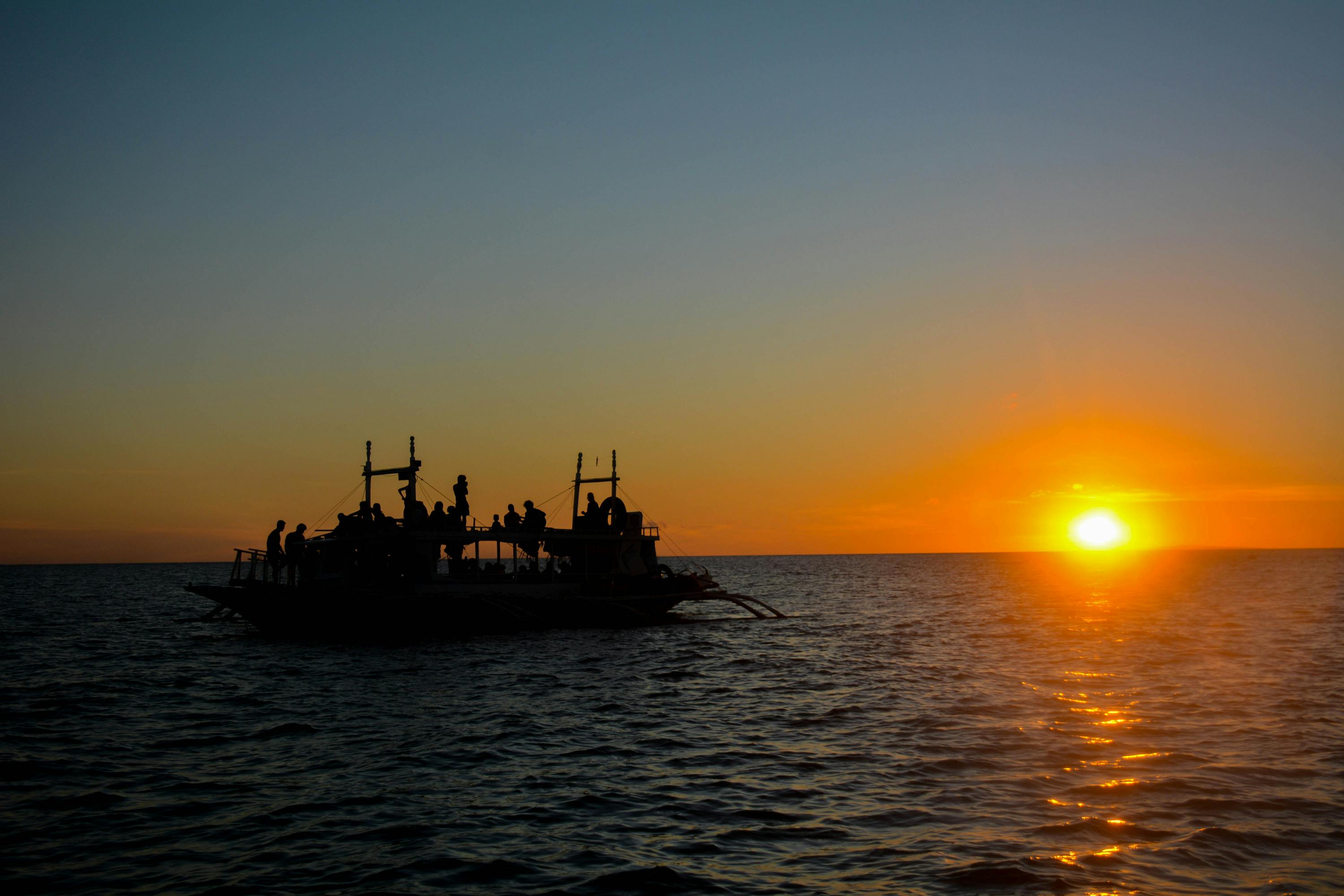 A silhouetted boat with people on it against a vibrant sunset over calm ocean water.