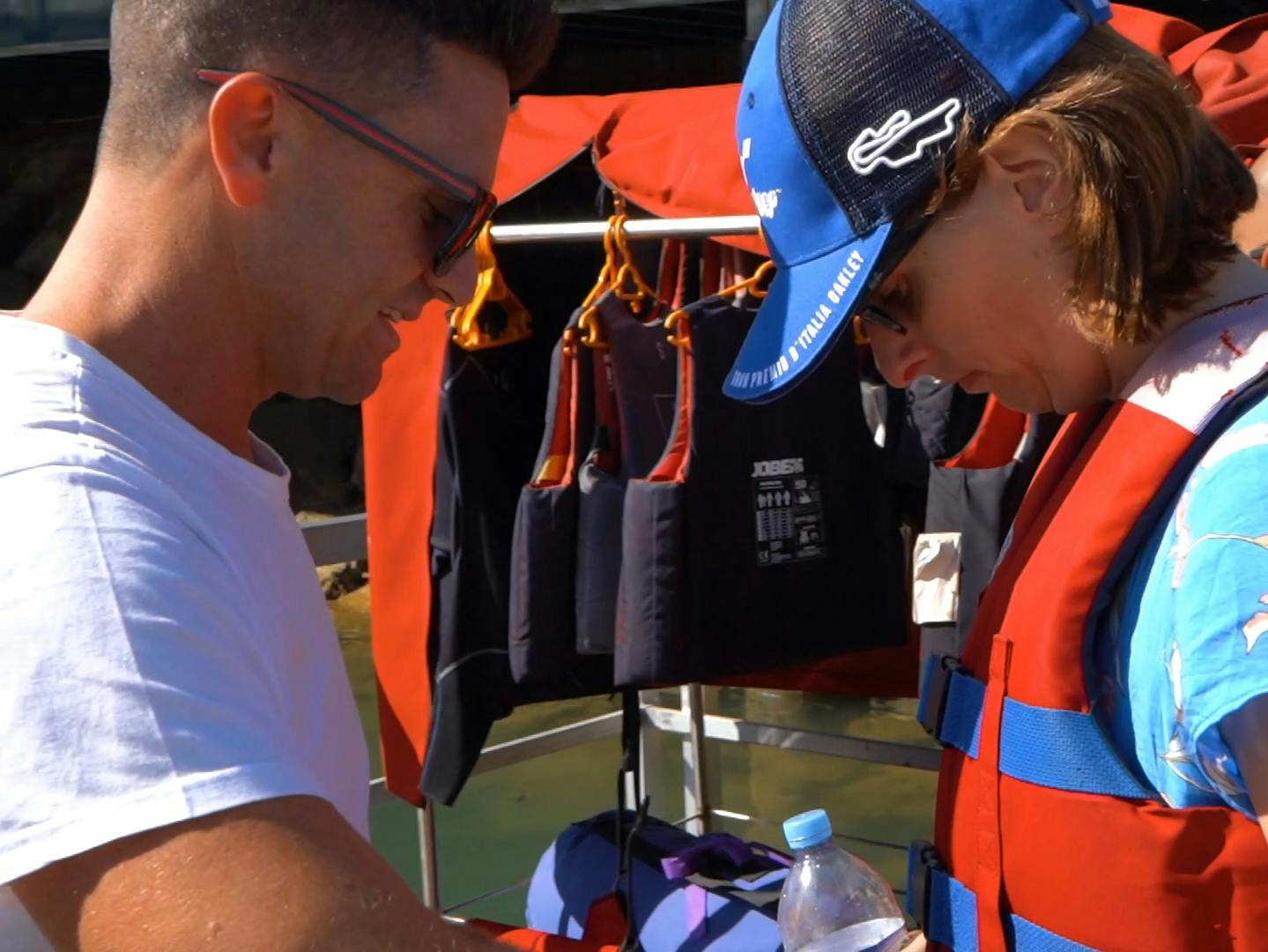 Dva lidé se připravují na vodní aktivitu a v blízkosti háčků mají na sobě záchranné vesty s větším počtem záchranných vest. Jedna osoba drží láhev na vodu.