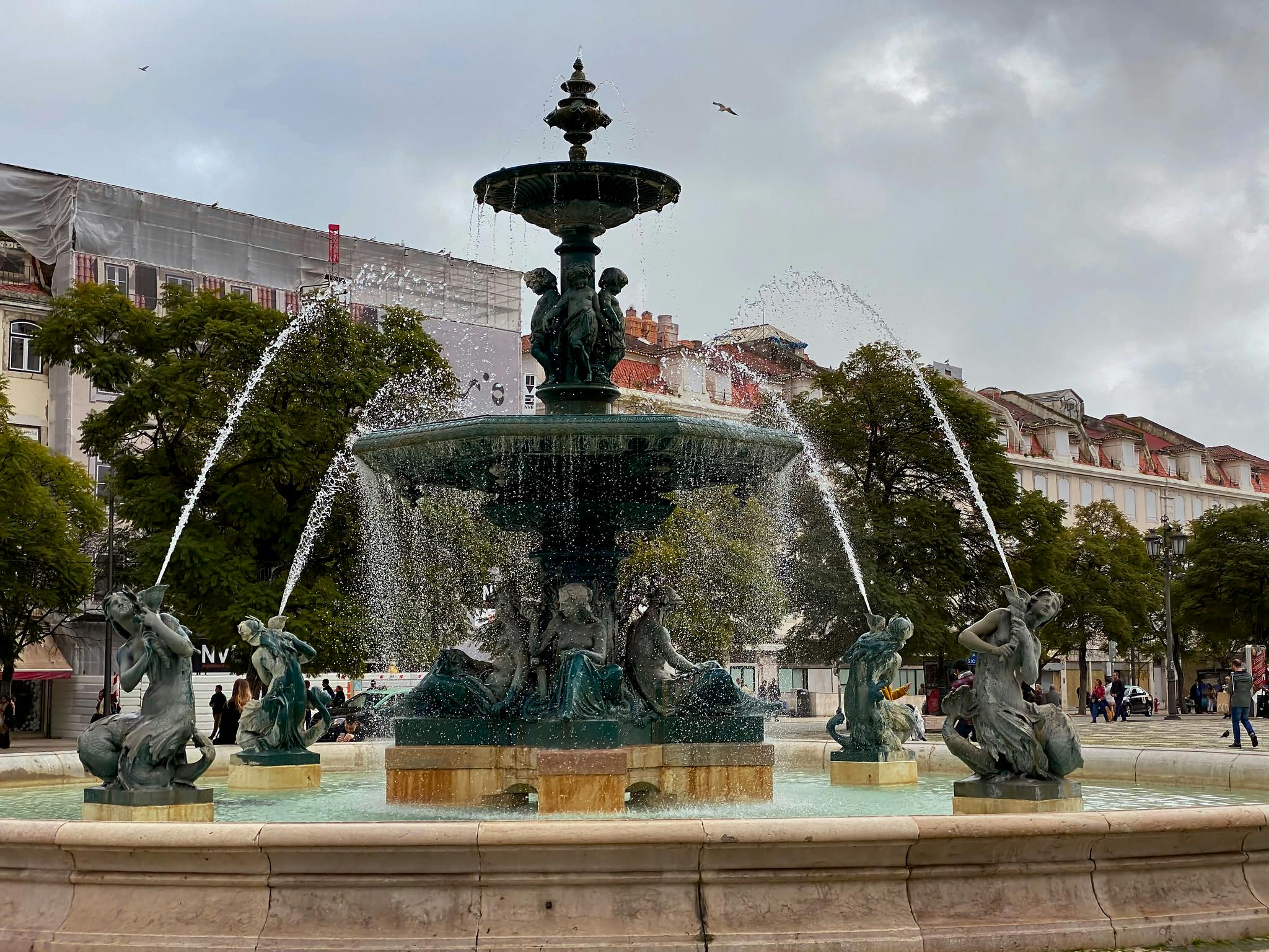 En dekorativ fontän med vattenpipar och statyer på ett offentligt torg, omgivet av träd och historiska byggnader.