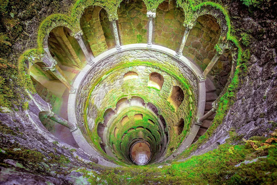 Circular, moss-covered stone well with multiple levels, arches, and columns, spiraling downward into the ground.