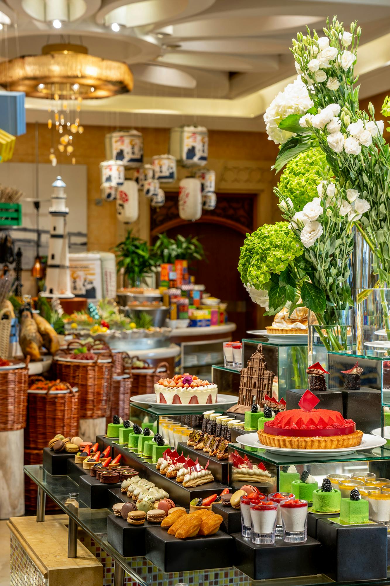 A vibrant display of colorful pastries and desserts on glass shelves, surrounded by flowers and decor in a bakery setting.