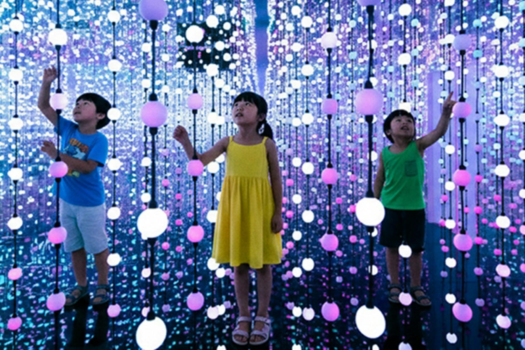 Two children stand among hanging strings of glowing orbs in a colorful, illuminated art installation.