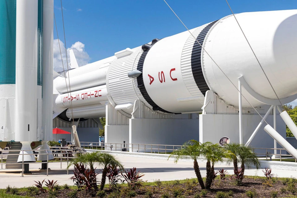 A large white rocket displayed outdoors on a sunny day, surrounded by plants and a paved walkway.