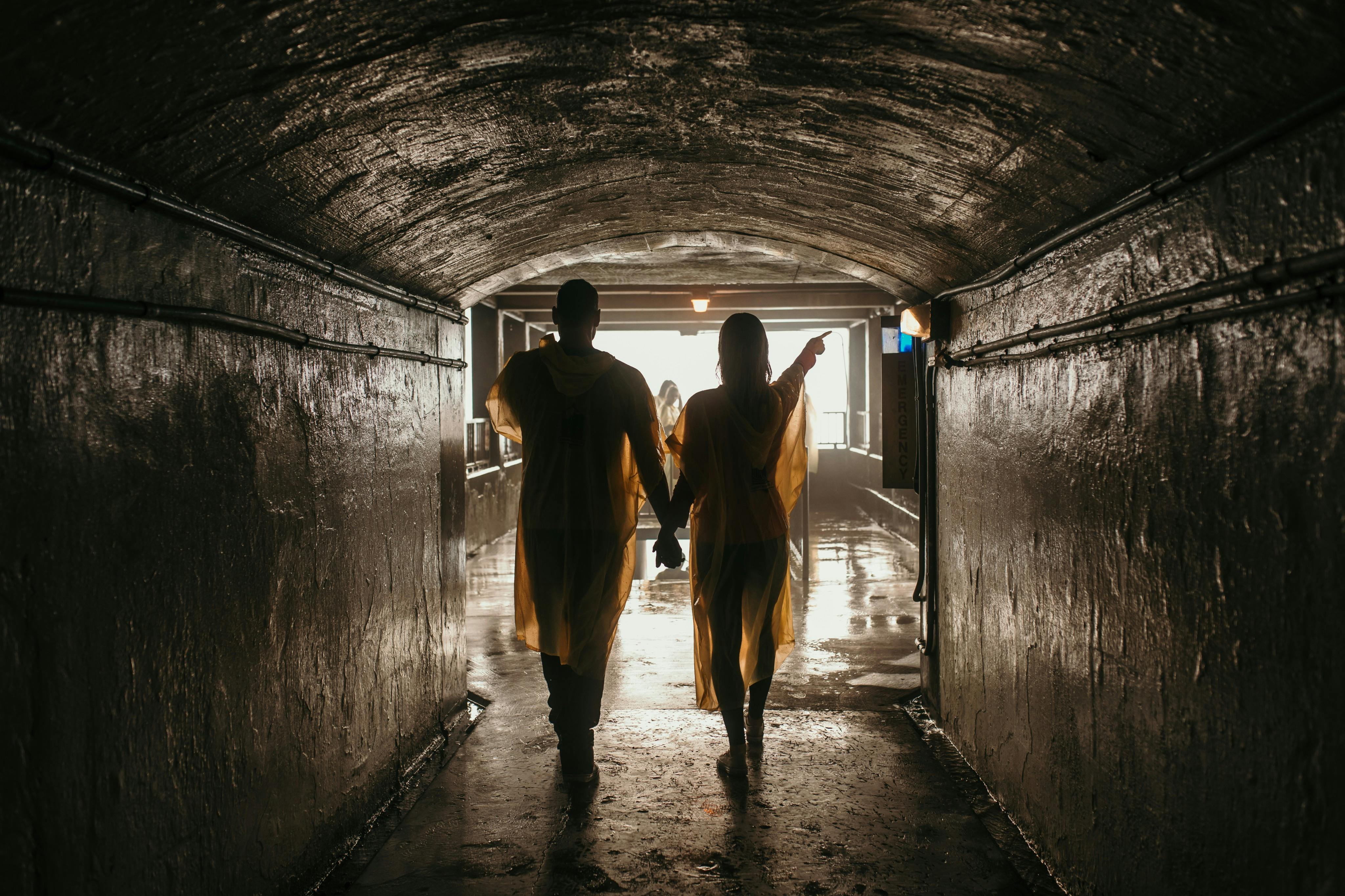 Dva lidé ve žlutých dešťových pončích jdou ruku v ruce slabě osvětleným tunelem k osvětlenému východu.