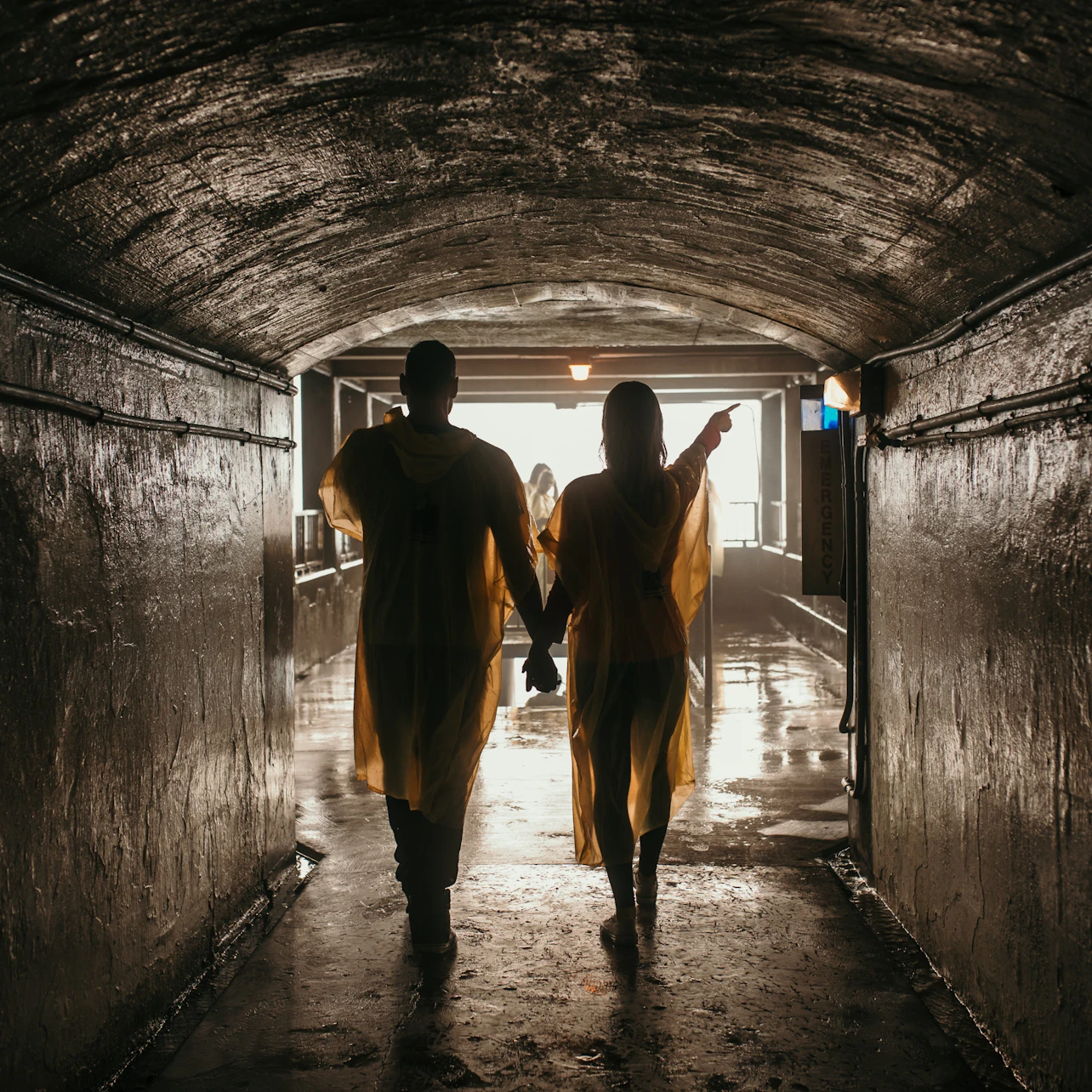 Two people wearing yellow rain ponchos walk hand in hand through a dimly lit tunnel towards an illuminated exit.
