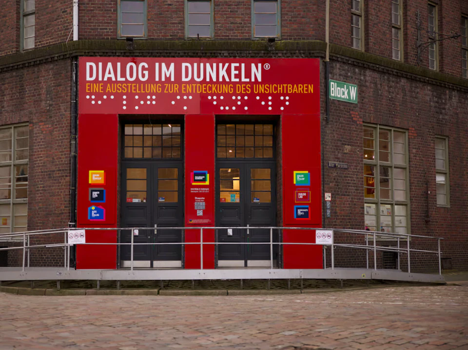 Entrance to “Dialog im Dunkeln” exhibition in a red-brick building with signs in German and a wheelchair ramp.