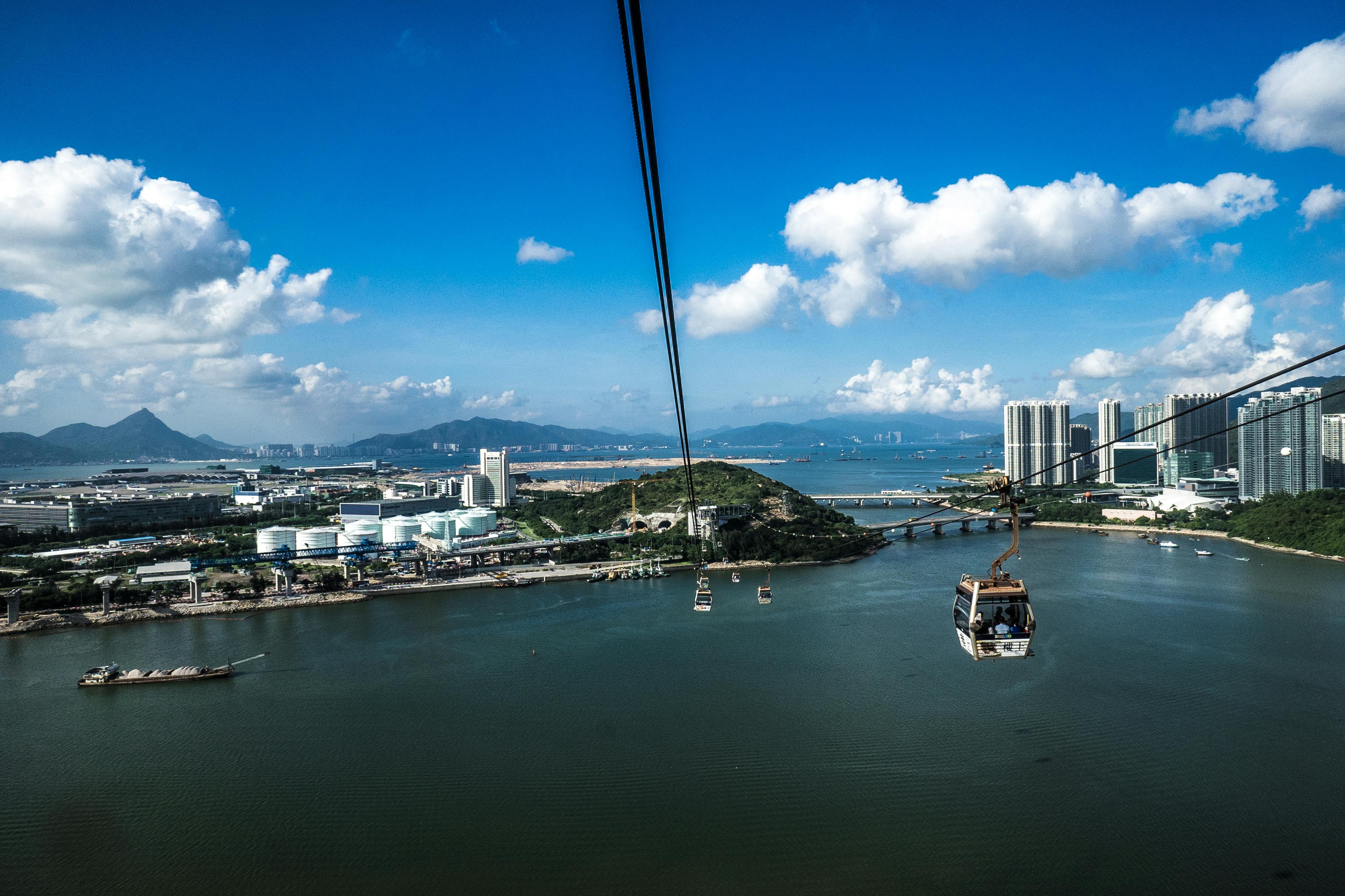 맑고 푸른 하늘 아래 건물과 언덕, 다리를 배경으로 물 위에 매달린 케이블카.