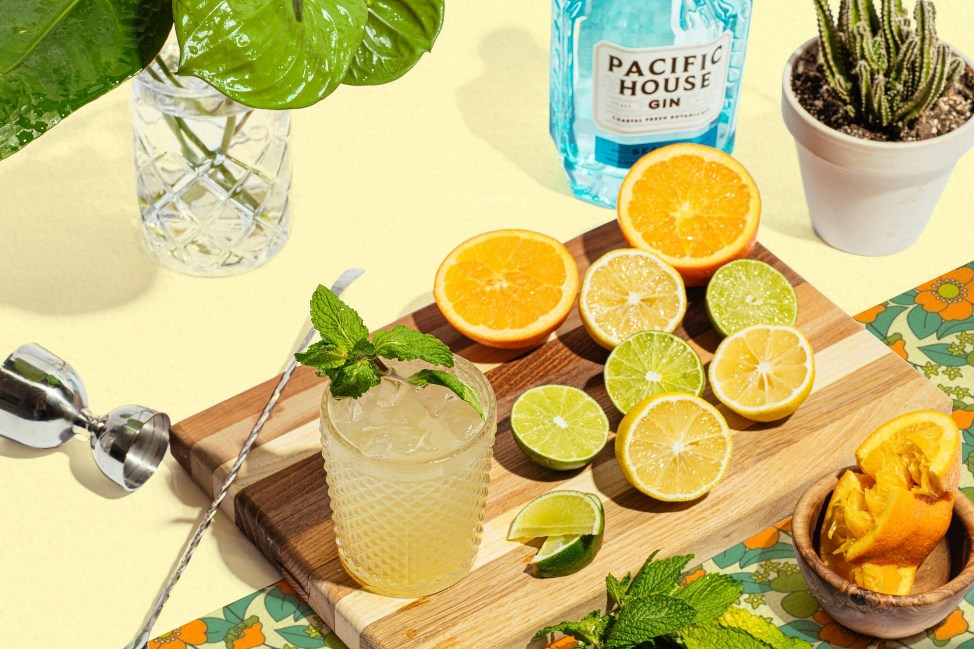 A wooden board with sliced citrus fruits, a glass with a cocktail and mint garnish, and a bottle of gin in the background.