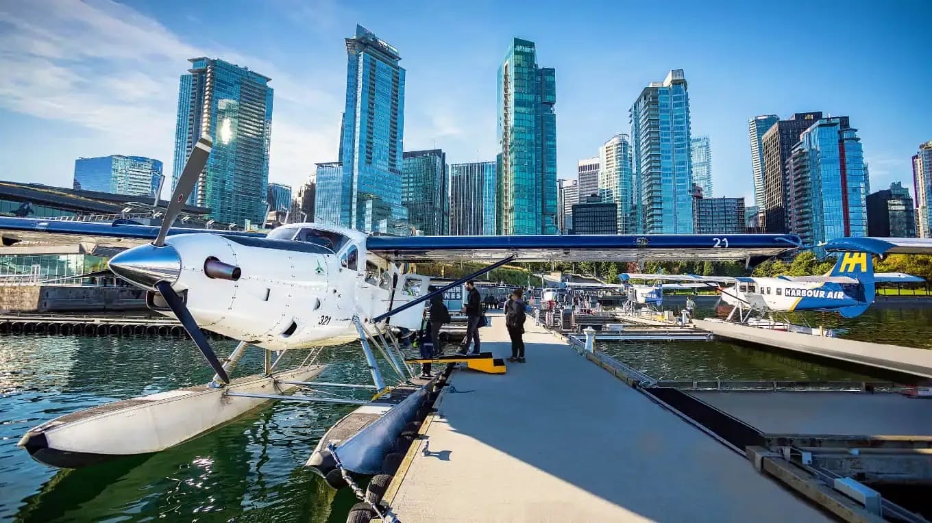 Zwei angedockte Wasserflugzeuge vor einer Stadtlandschaft am Wasser mit hohen Gebäuden unter blauem Himmel. Menschen stehen auf dem Pier.