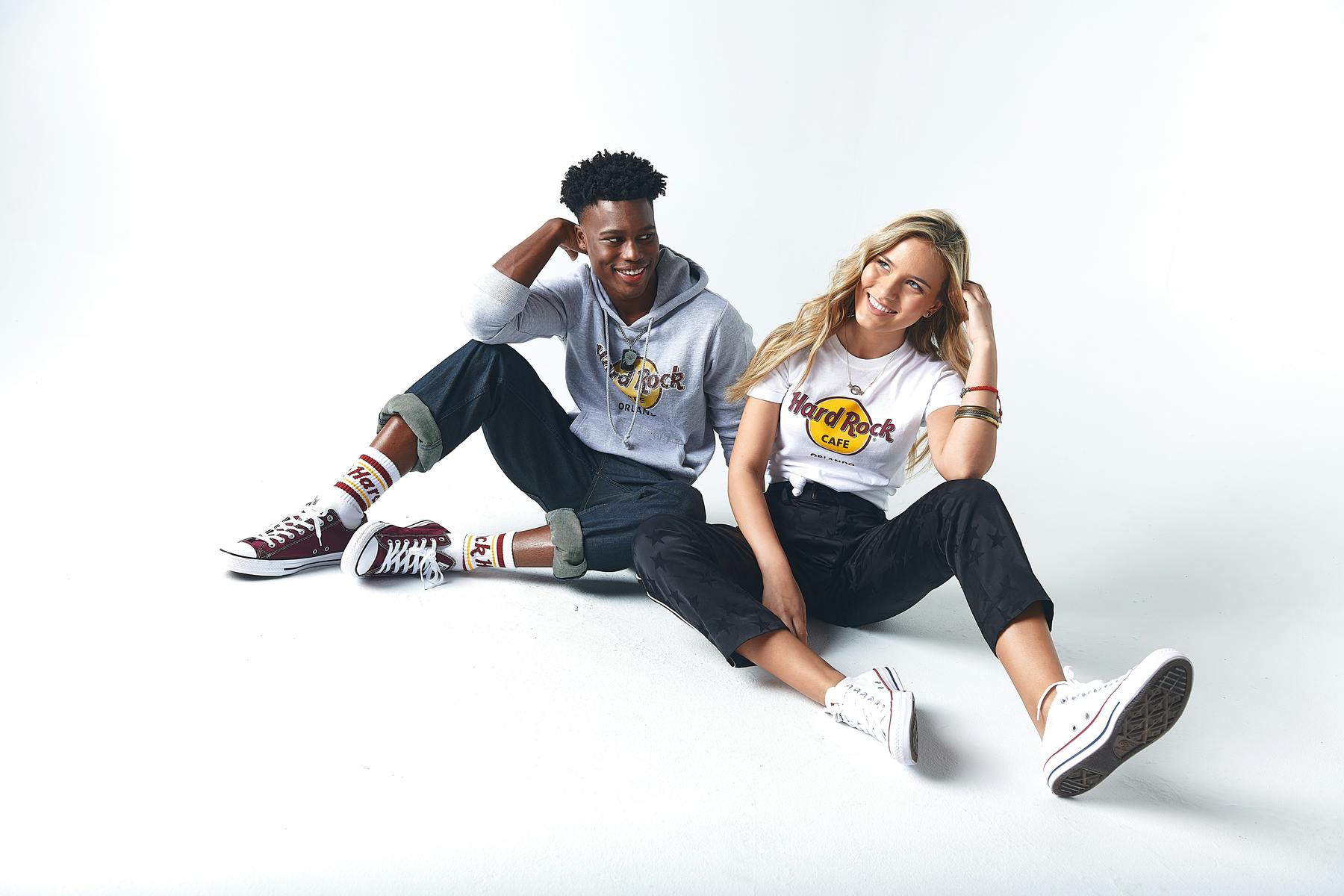 A man and woman sit on the floor wearing Hard Rock Cafe clothing and Converse sneakers, smiling against a white background.
