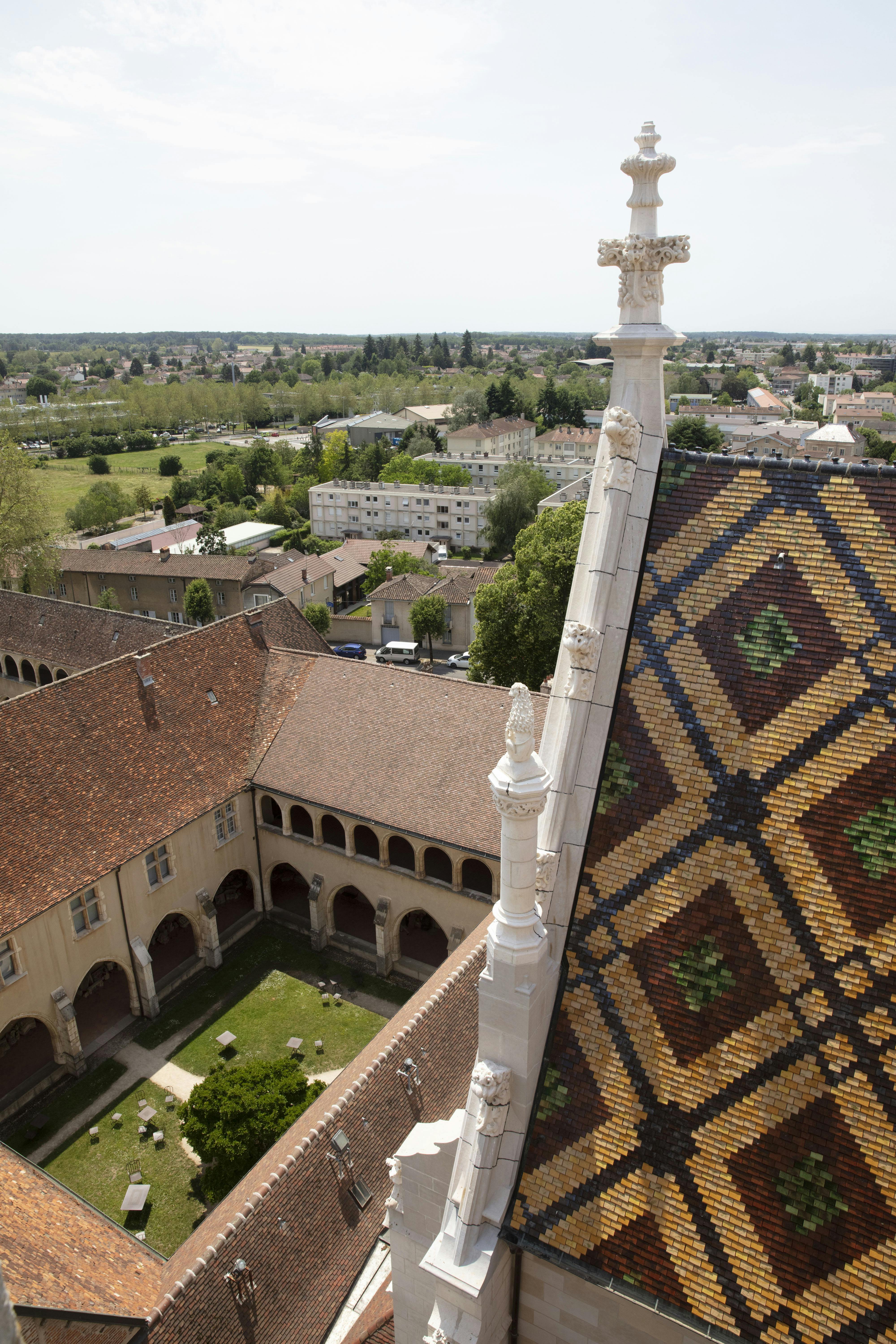 Colorful tiled roof of a historic building, overlooking a peaceful courtyard with arches, and a town with greenery in the background.