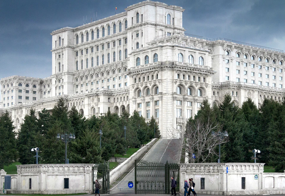 Large neoclassical building with ornate details, surrounded by trees, and a staircase leading up from a gated entrance. People walk below.