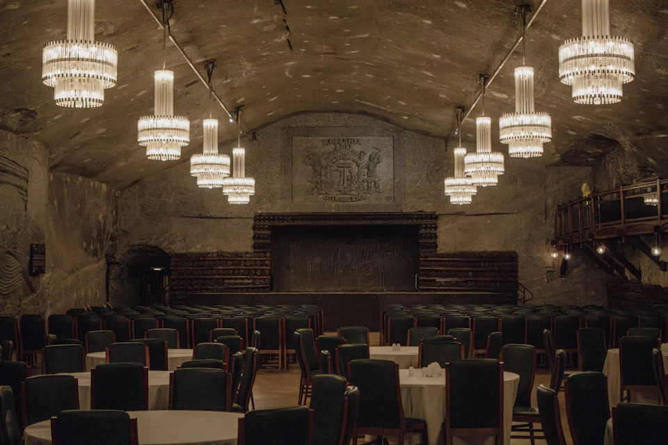 A grand dining hall with chandeliers, stone walls, a carved mural, and rows of tables and chairs in an elegant setting.