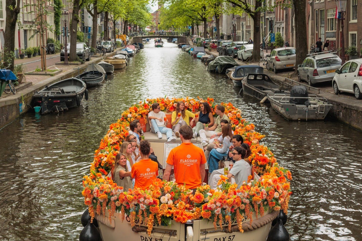 A boat decorated with orange flowers carries people along a canal, surrounded by parked cars and trees on a sunny day.