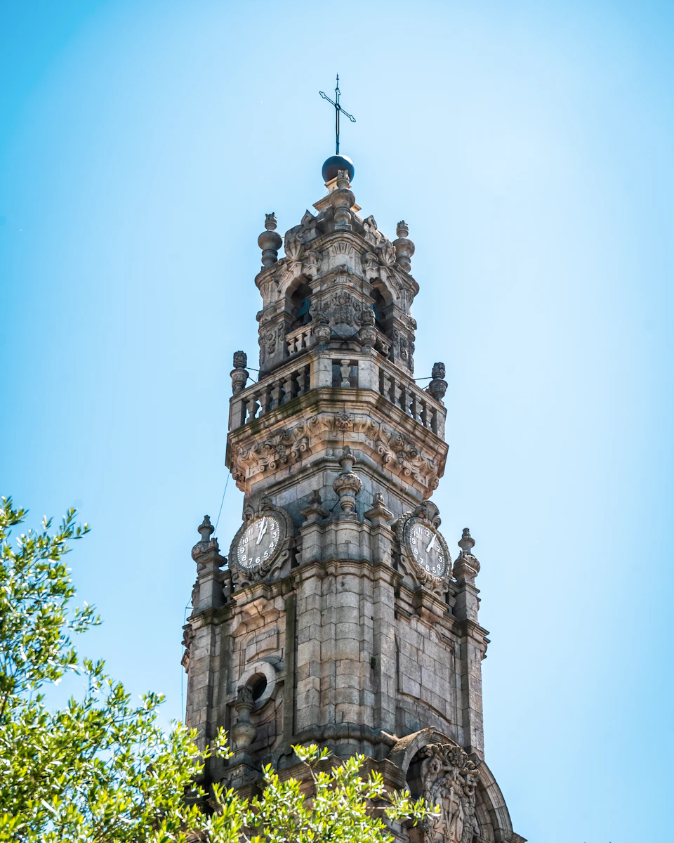 A historic stone clock tower with ornate details under a clear blue sky, with tree branches visible in the foreground.