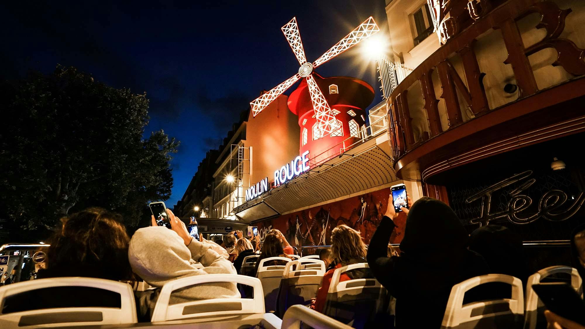 Ludzie w autobusie robią nocą zdjęcia oświetlonego wiatraka Moulin Rouge.