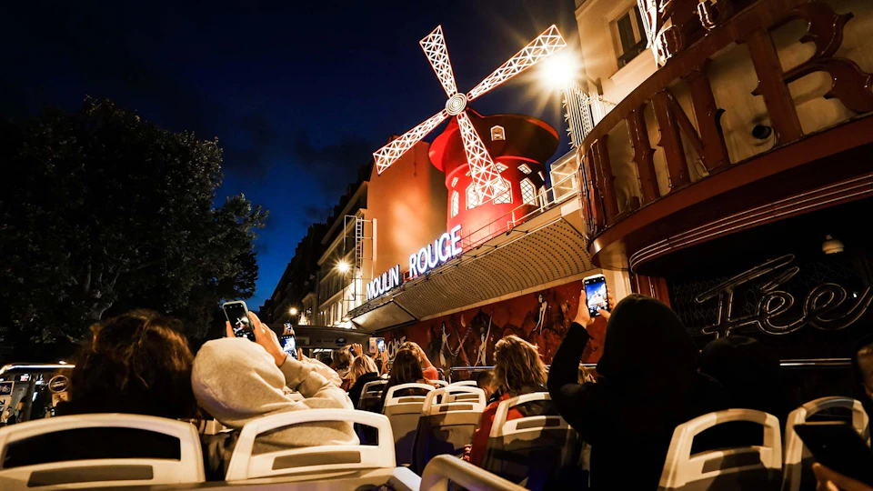 People on a bus taking photos of the illuminated Moulin Rouge windmill at night.