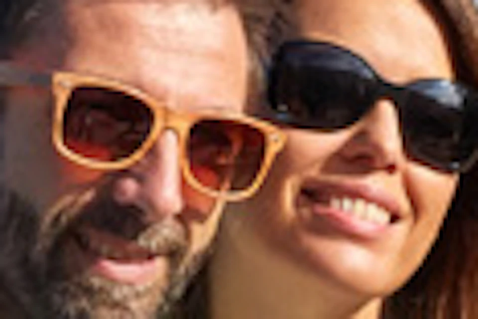 A couple sits on a rocky ledge at the beach, smiling while posing for a photo, with buildings and the ocean in the background.