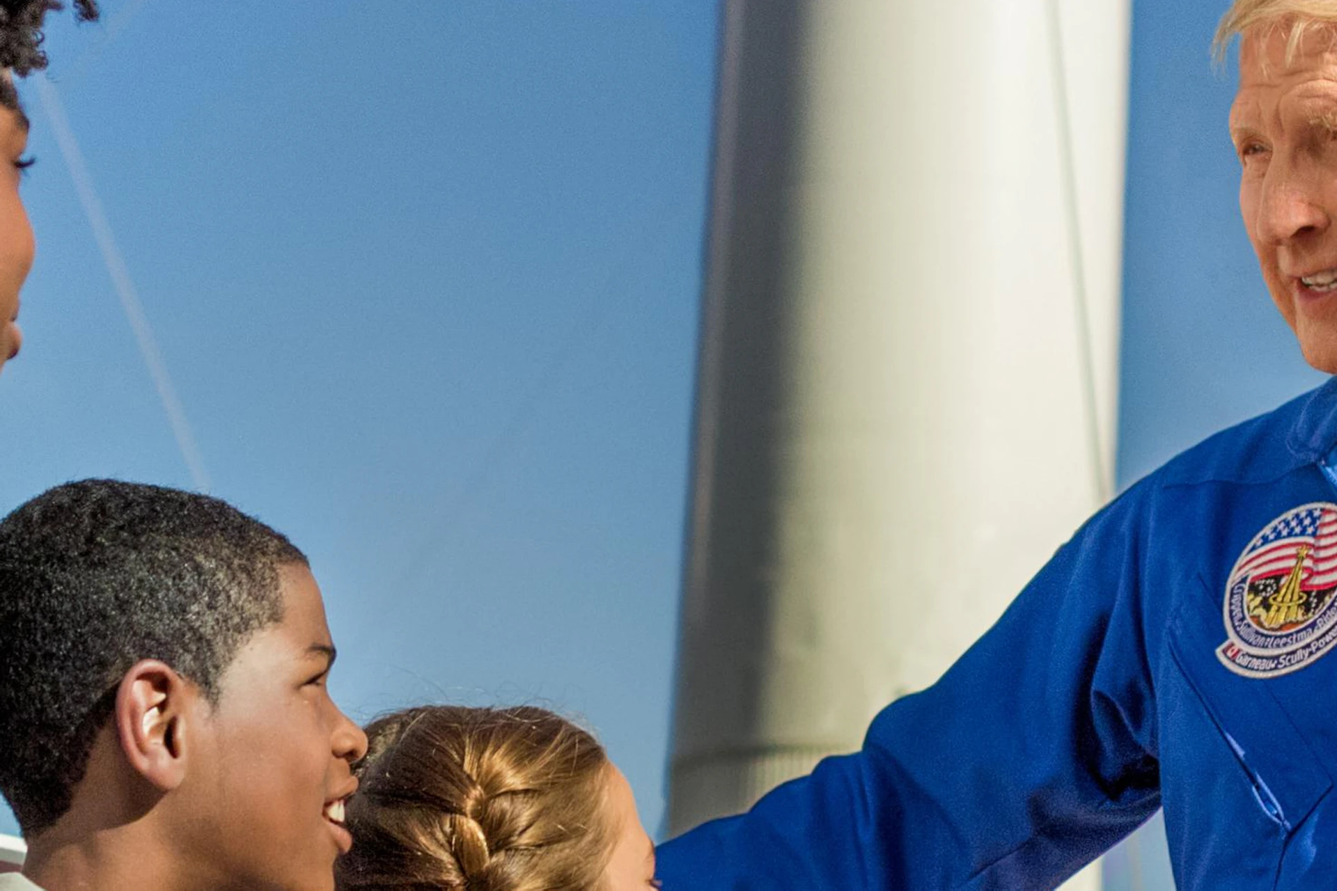 A man in a blue NASA spacesuit talking to a group of children with rockets in the background on a sunny day.