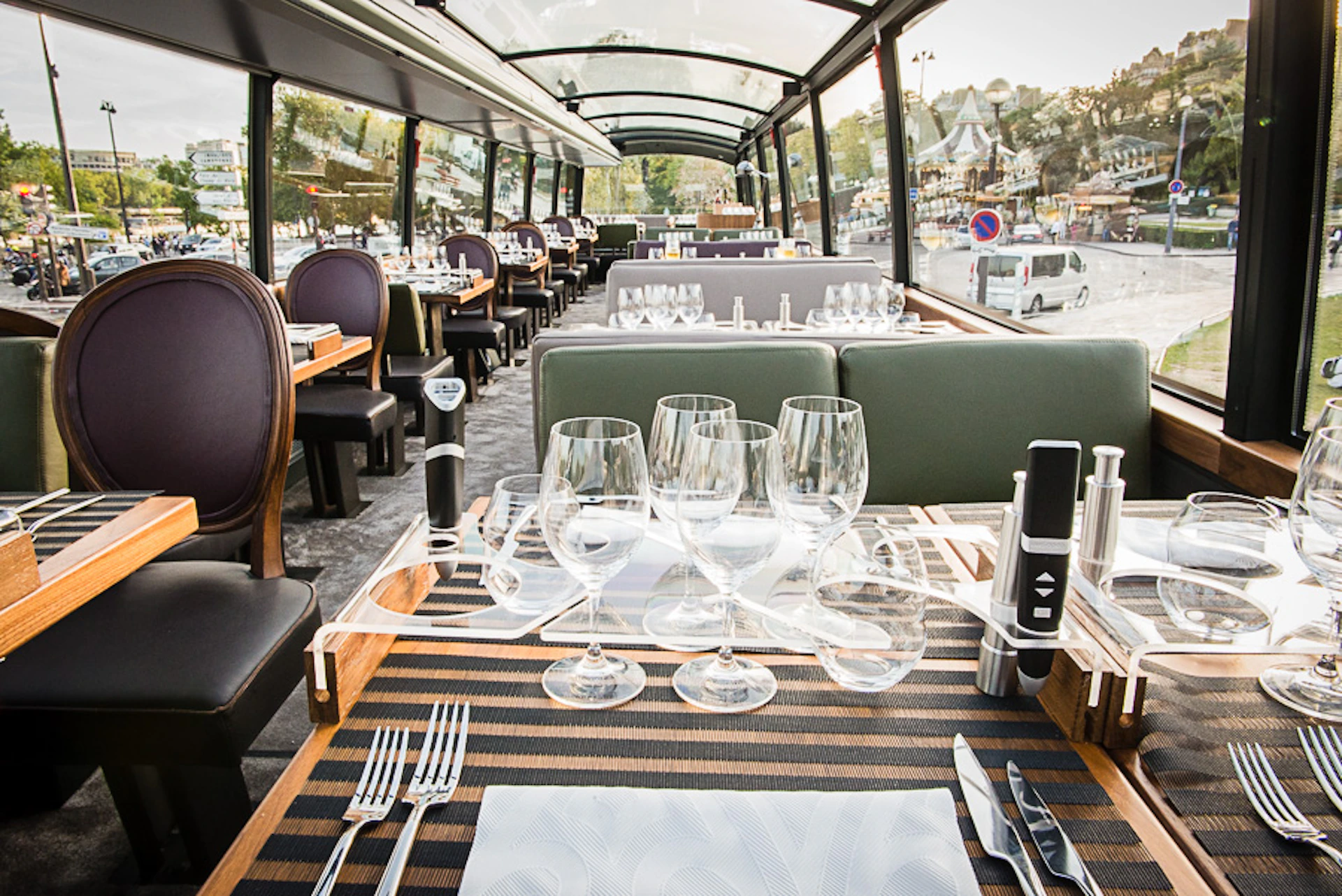 An elegantly set dining table with empty wine glasses and cutlery inside a glass-roofed restaurant bus with exterior city views.
