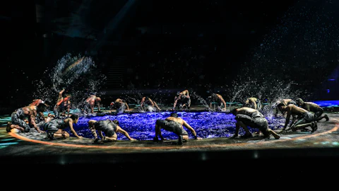 Performers in black outfits splashing water on stage under spotlights, surrounded by darkness in the background.