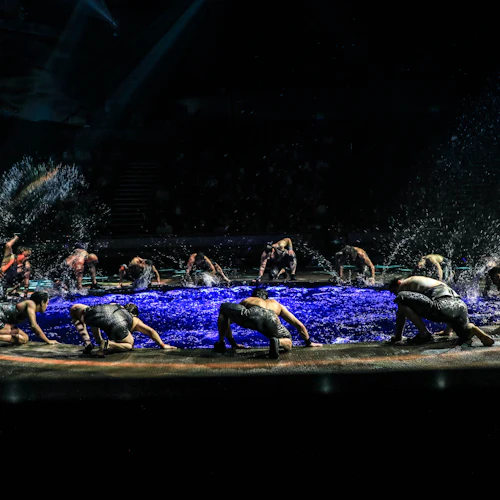 Performers in black outfits splashing water on stage under spotlights, surrounded by darkness in the background.