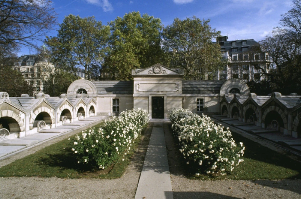 Stone mausoleum with arched tombs surrounded by blooming white flowers, pathways, and trees under a clear blue sky.