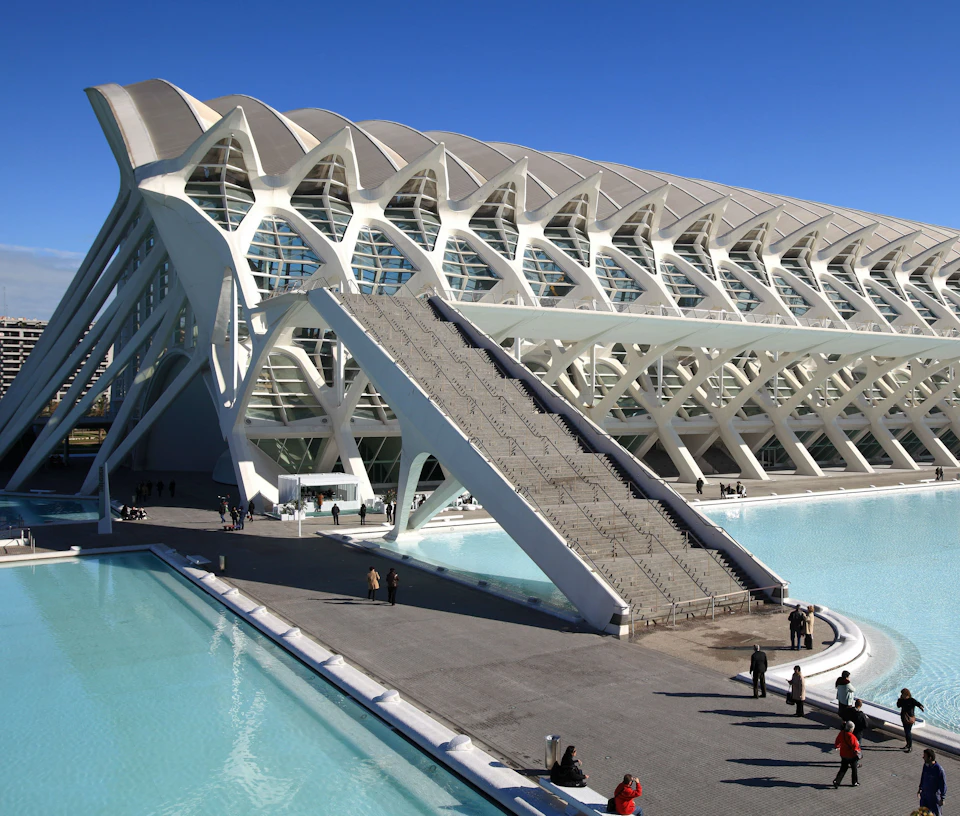 Futuristic building with an intricate white lattice structure, adjacent to a large reflective pool, under a clear blue sky.