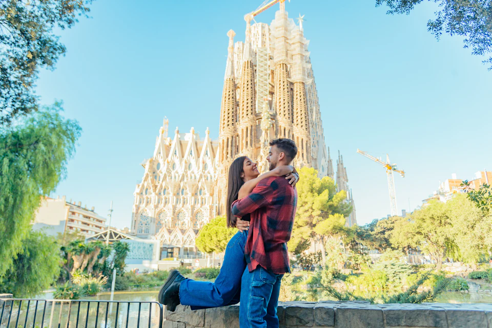 A couple embraces joyfully in front of the Sagrada Família with a clear sky and cranes in the background.