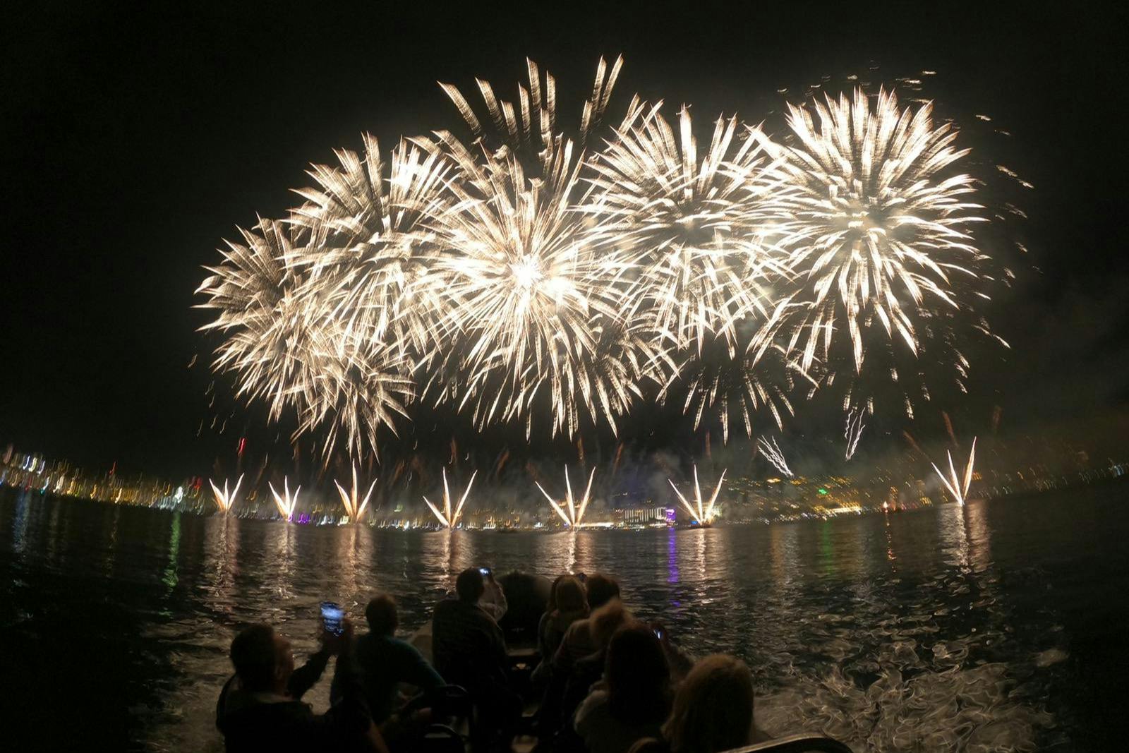 People on a boat watching large, bright fireworks explode in the night sky over a city skyline reflecting on the water.