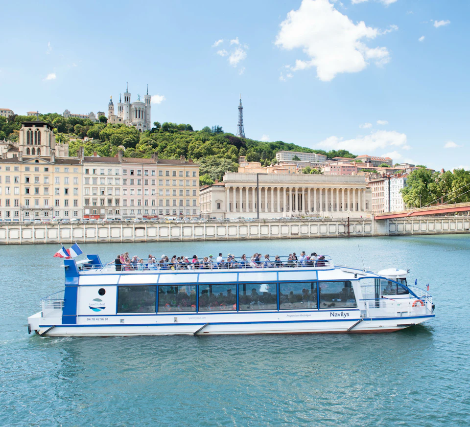 Tourist boat with an open top deck on a river, with historic buildings and a basilica on a hill in the background on a clear day.