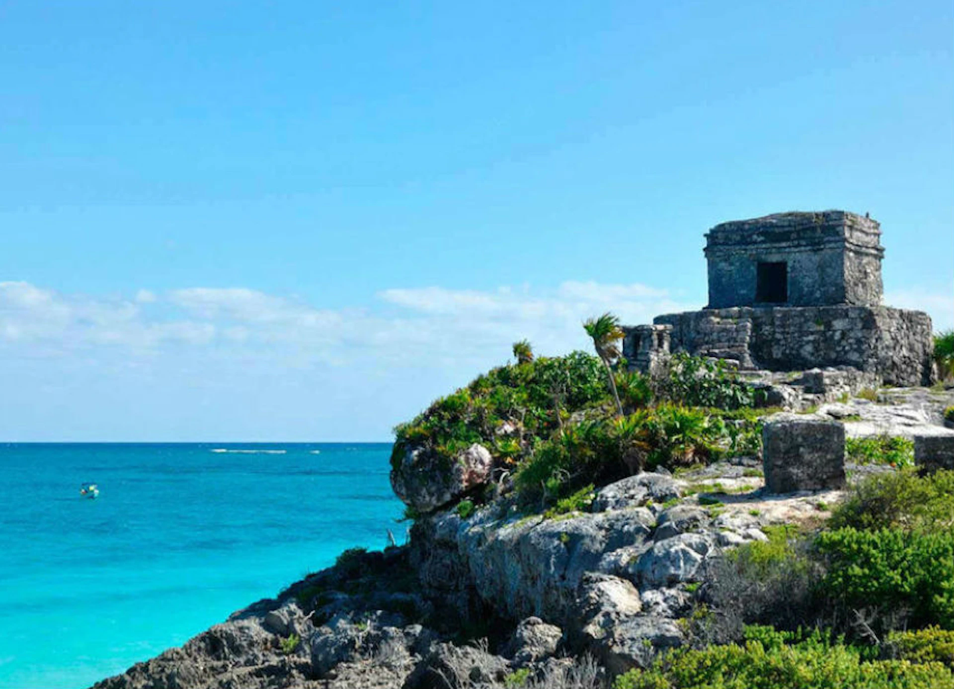 Ancient stone ruins sit atop a rocky cliff overlooking a clear blue ocean under a bright, cloud-dotted sky.