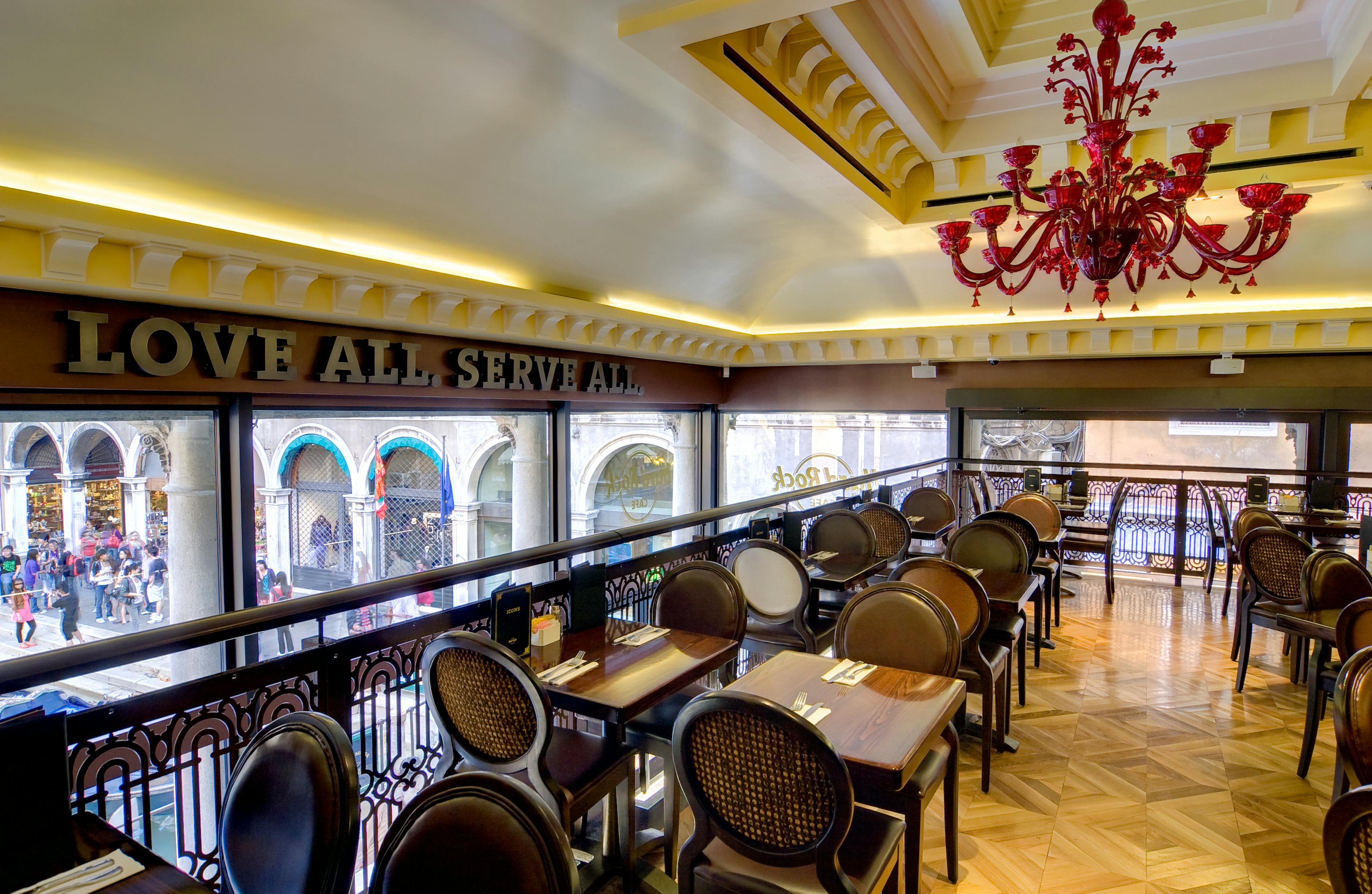 Modern indoor dining area with brown chairs, wooden tables, a red chandelier, and large windows overlooking an architectural street view.