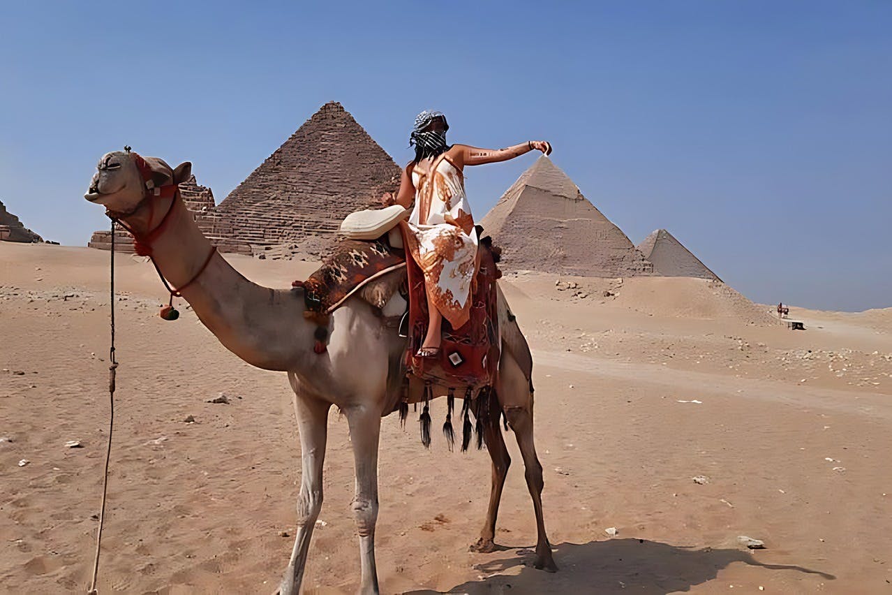 在晴朗的蓝天下，一个人在沙漠景观的金字塔附近骑着骆驼。