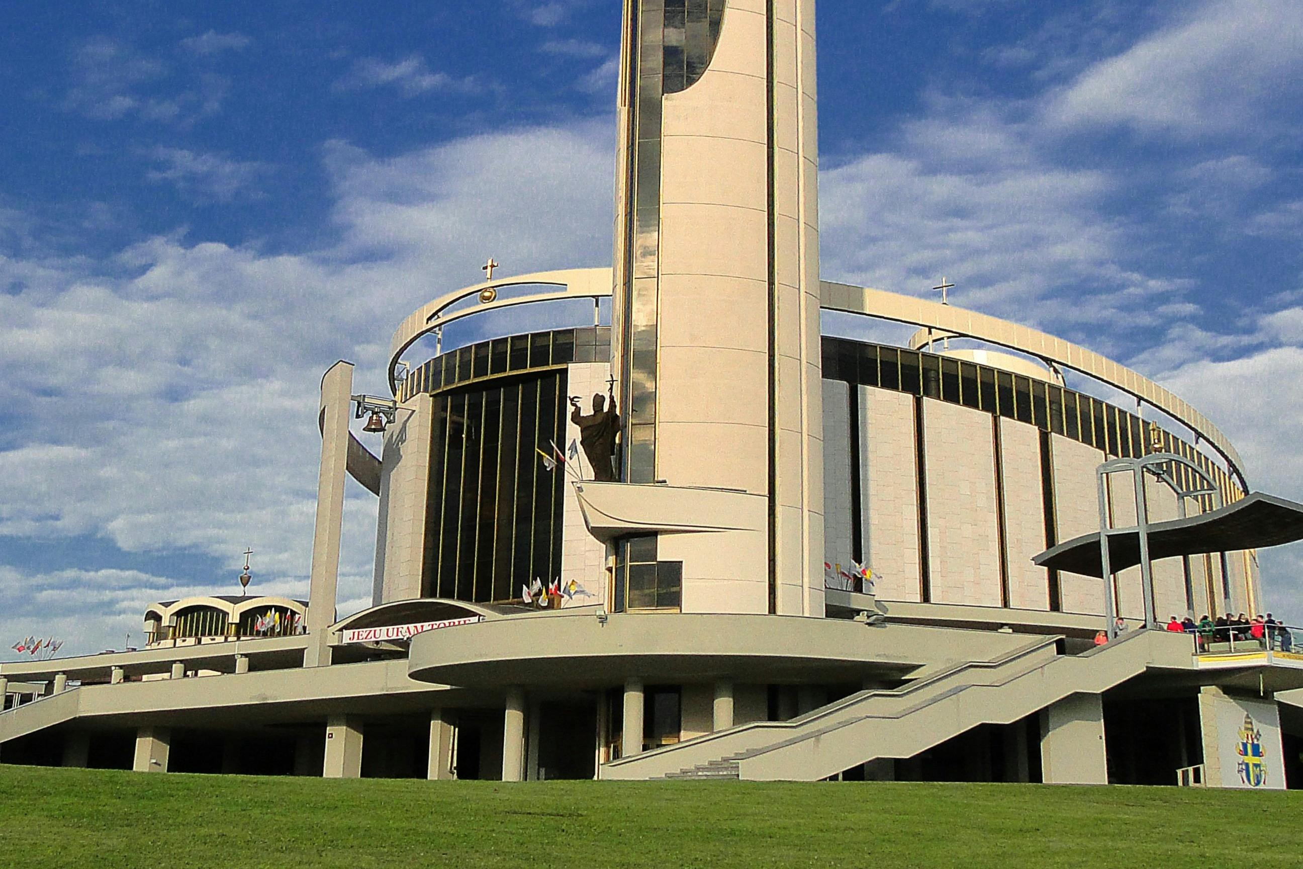 En modern kyrka med en cylindrisk struktur, kors på toppen, en stor staty och trappor som leder till flera ingångar.