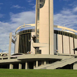 En modern kyrka med en cylindrisk struktur, kors på toppen, en stor staty och trappor som leder till flera ingångar.