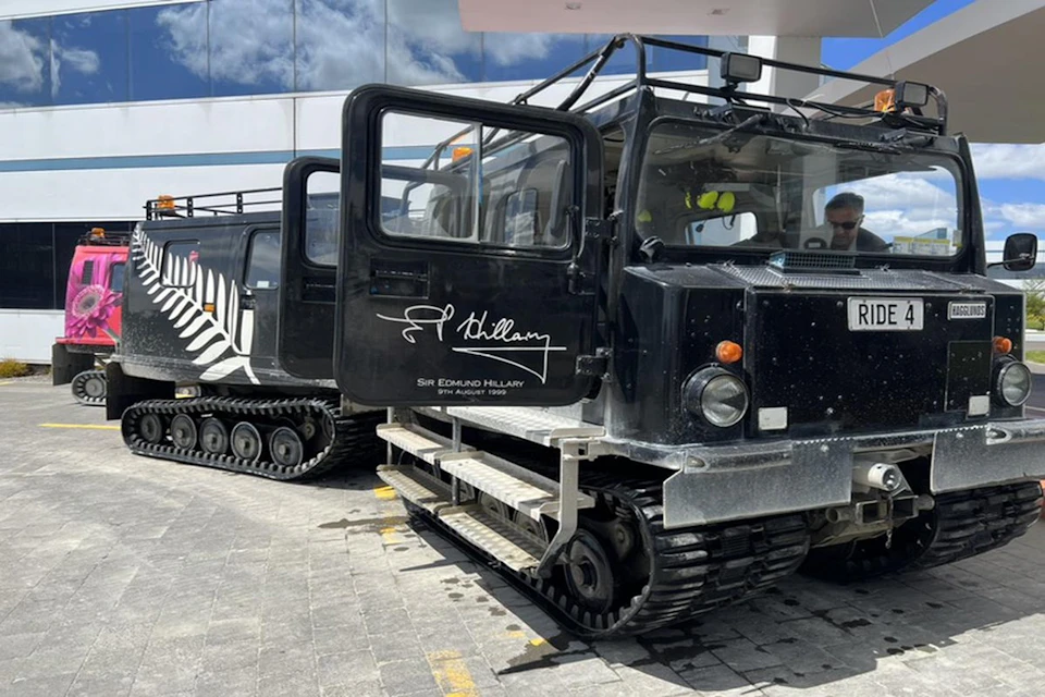 A tracked vehicle with "Sir Edmund Hillary" signature on its side is parked on a paved area under a partly cloudy sky.