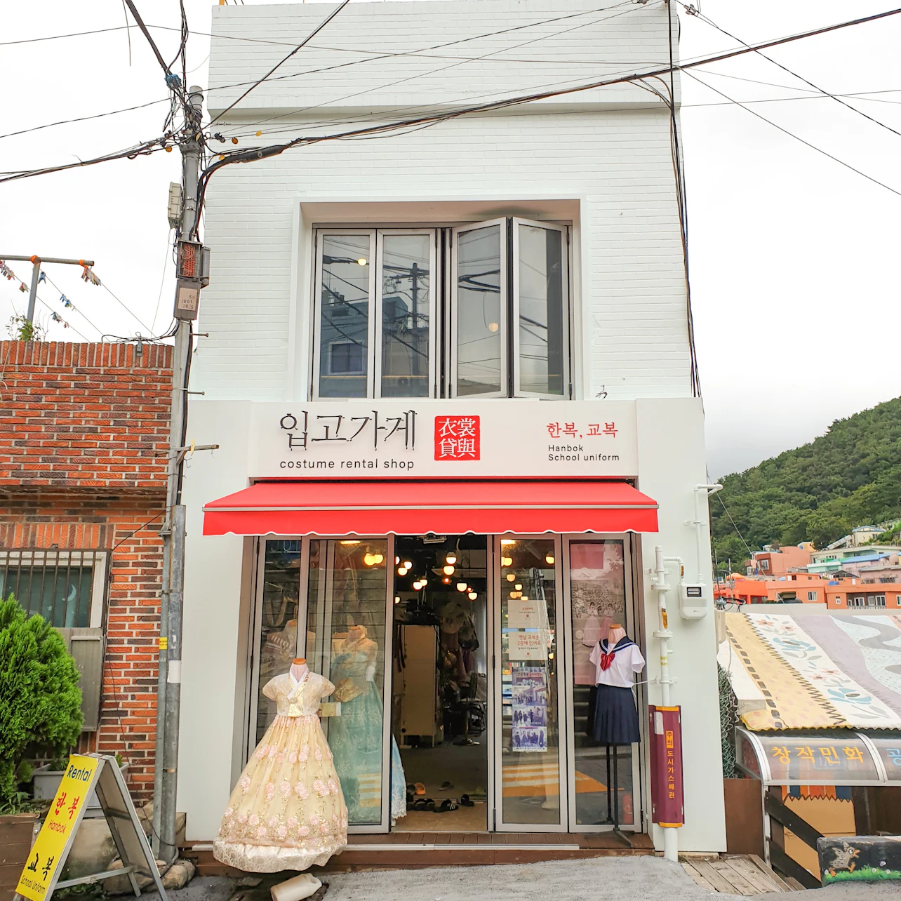 A two-story building with a signage indicating a costume rental shop. Dresses are displayed at the entrance under a red awning.