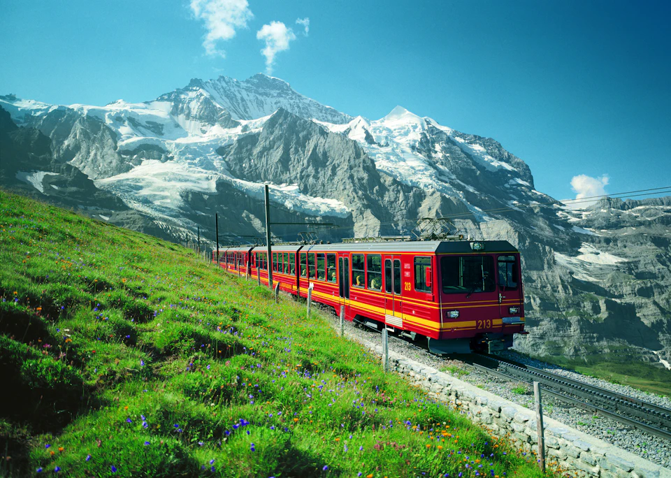 A red and yellow mountain train traveling up a grassy slope with snowy mountains in the background.