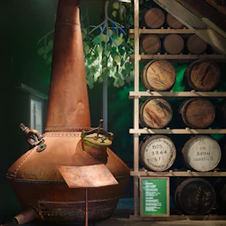 En stor koppardestillation fortfarande bredvid träfat på hyllor, med en lummig dekoration och en liten informationsplatta nedan.